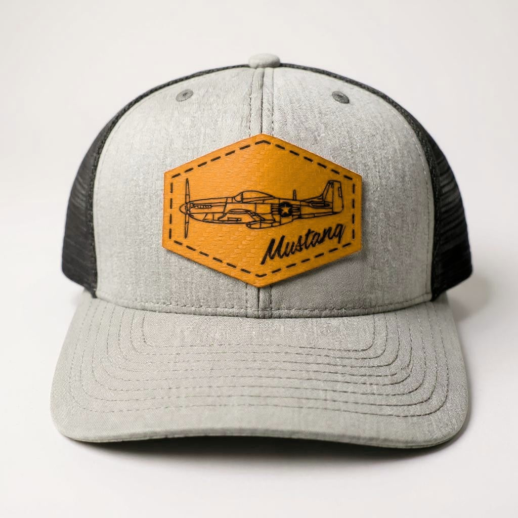 Gray and black trucker hat with a gold patch featuring an airplane and 'Mustang' text on a white background.